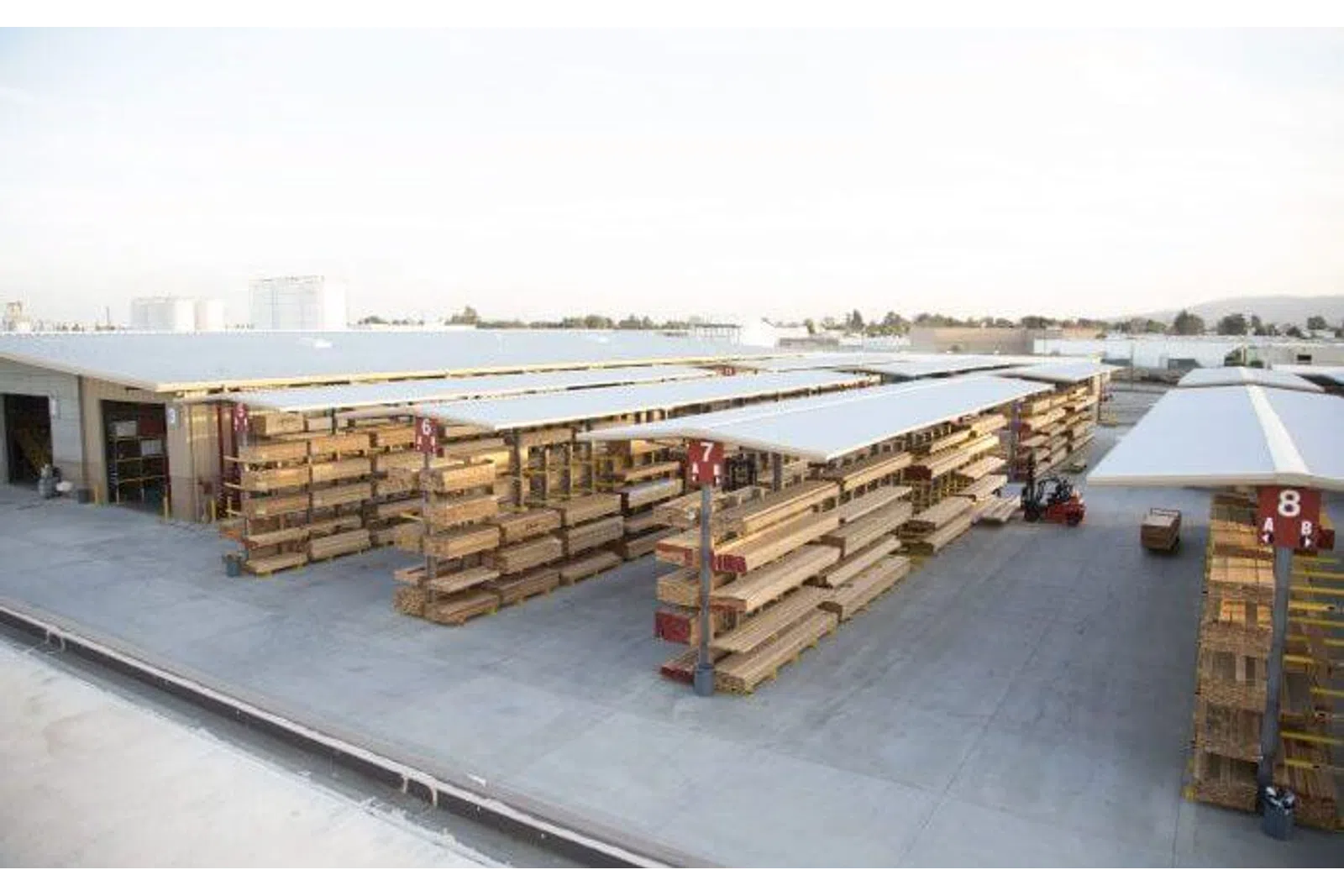 Outdoor lumber yard with organized stacks of wooden planks under white canopies and a forklift in use.