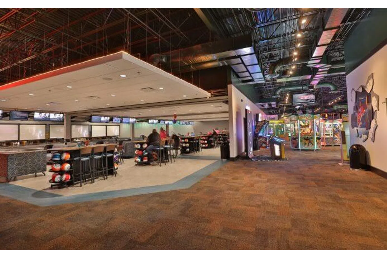 Spacious modern bowling alley with seating area, bowling balls, and arcade games under industrial ceiling.
