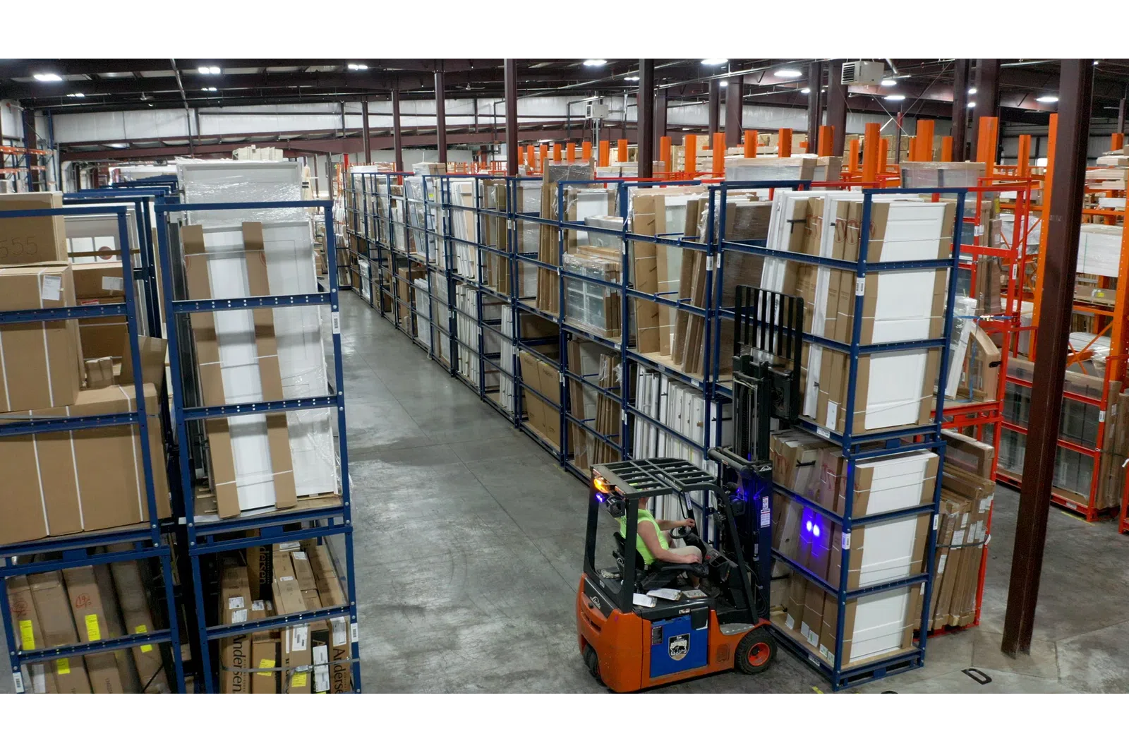 Forklift operator moving stacked boxes in a large organized warehouse with tall metal shelving units.