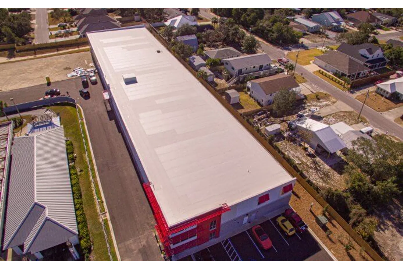 Aerial view of a large warehouse with a white roof and red accents surrounded by residential houses and parked cars.