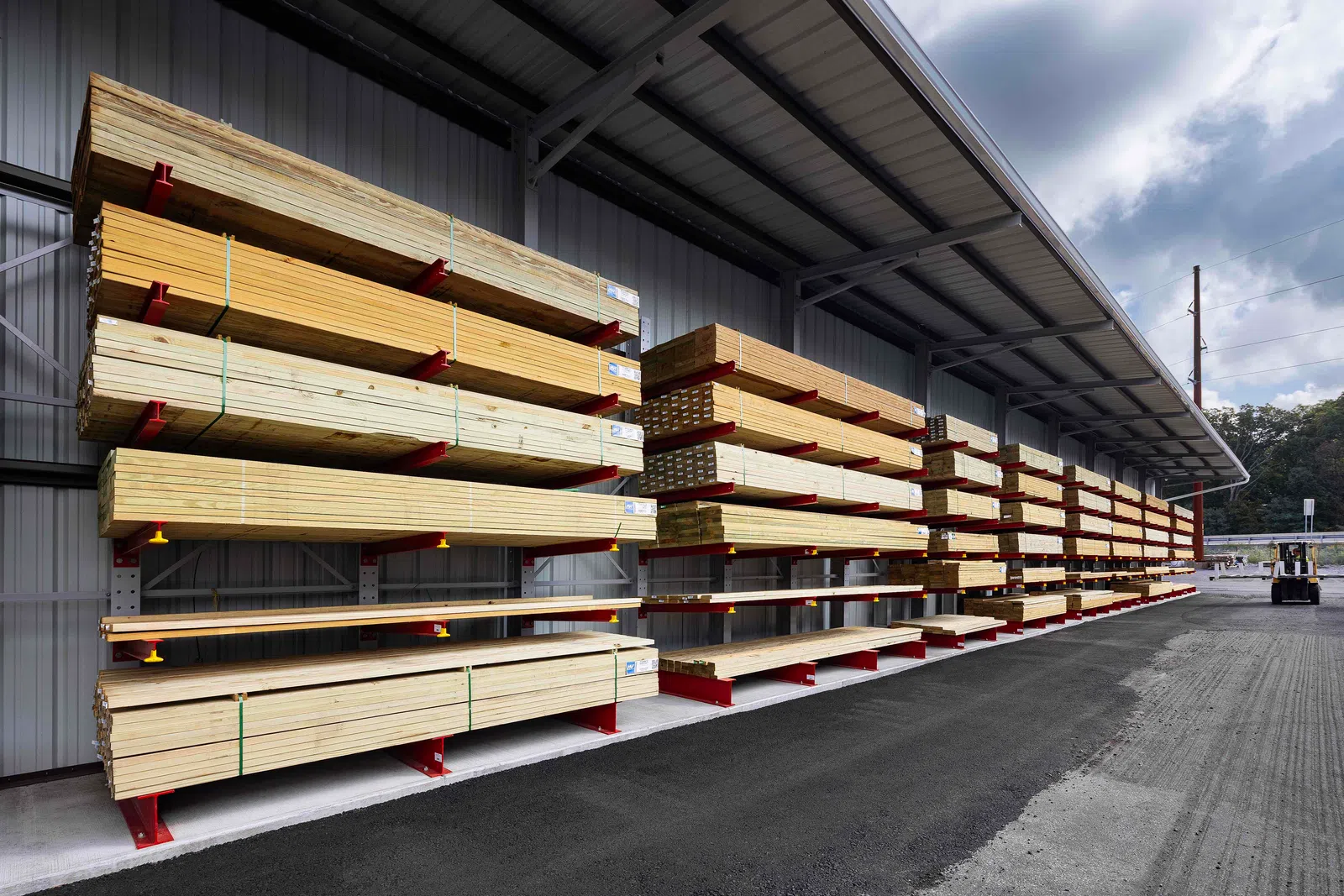 Outdoor lumber storage racks filled with stacked wooden planks under a metal canopy on a paved surface.