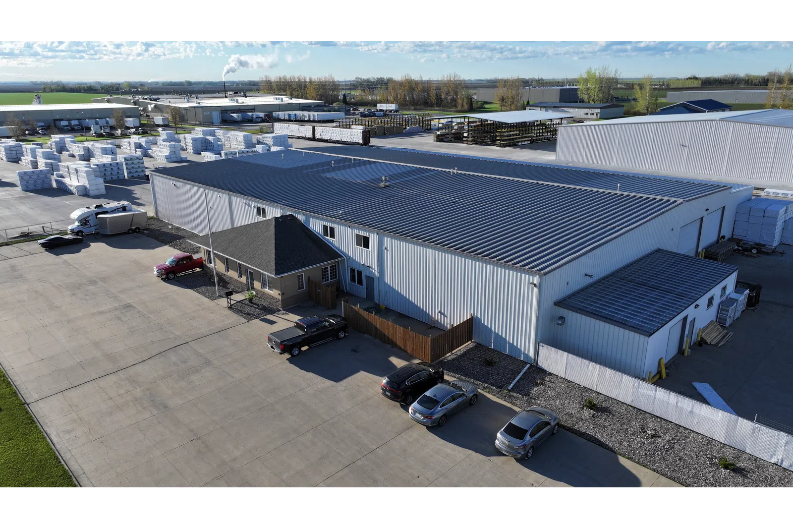 Aerial view of a large industrial warehouse with parked cars and stacks of materials outside under a partly cloudy sky