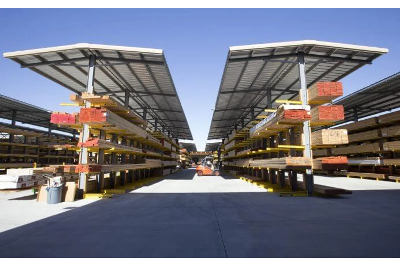 Outdoor lumber yard with organized stacks of wood under metal canopies and a forklift in the center aisle.