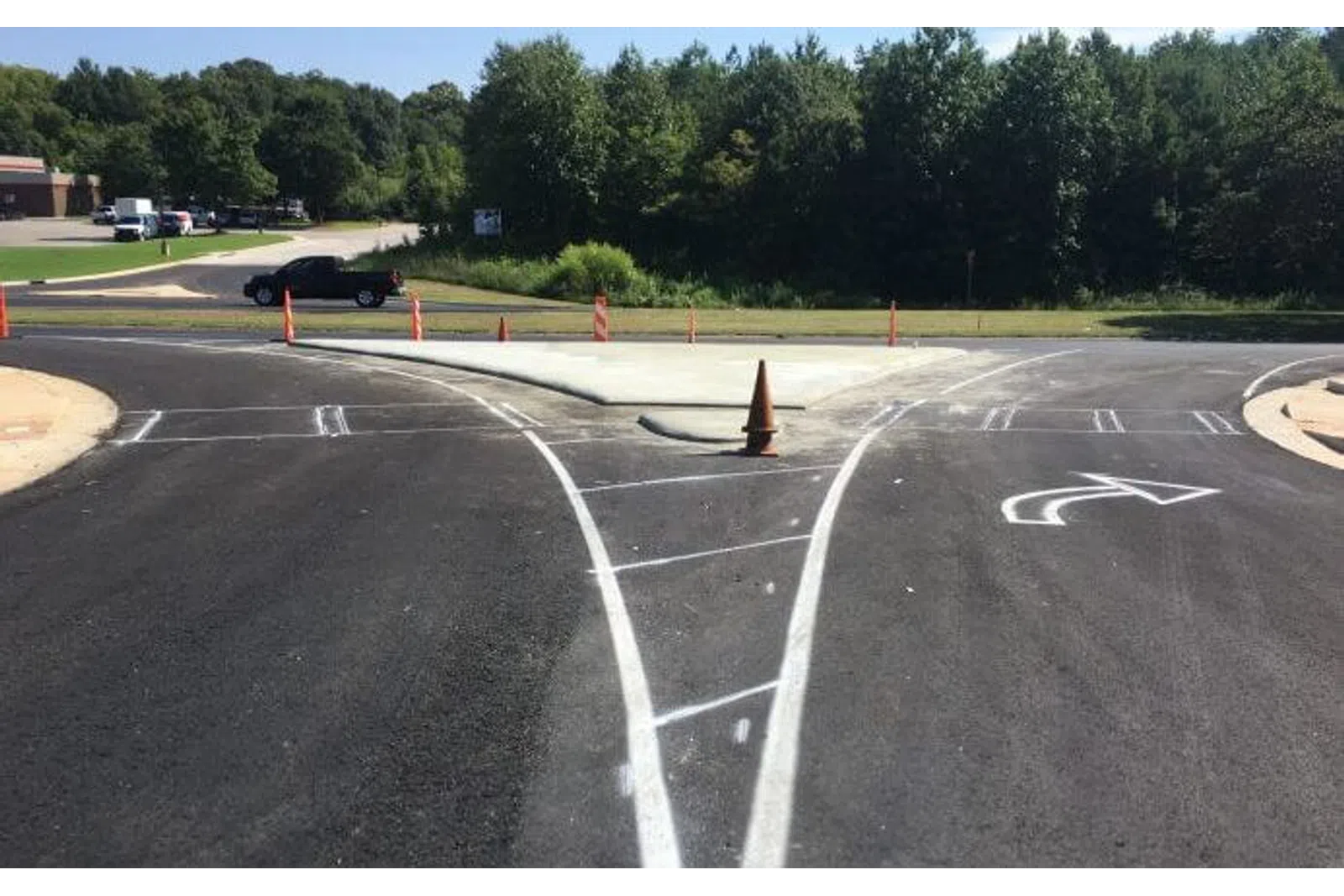 Freshly paved road with new white lane markings and a traffic cone at a fork under clear skies with trees.