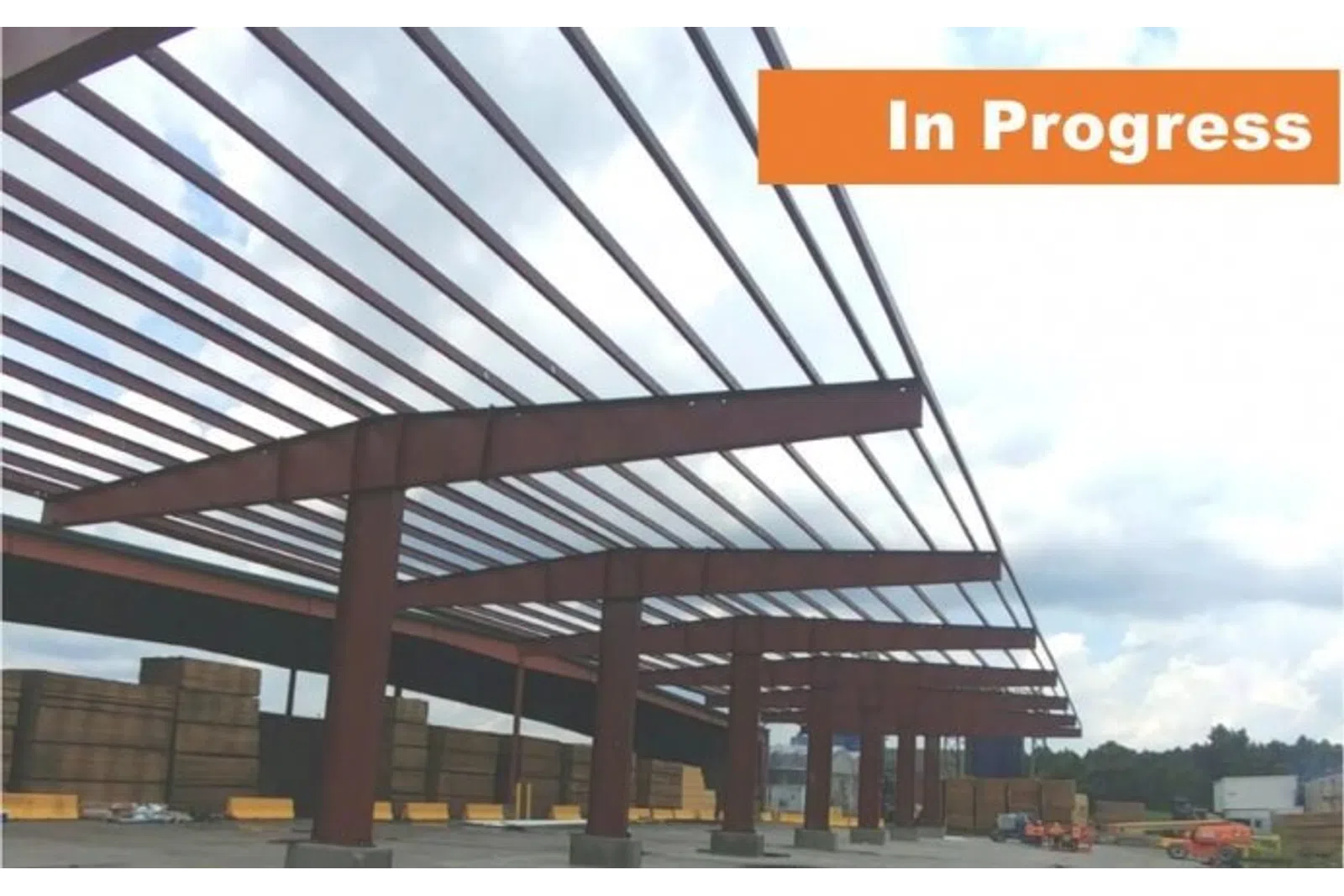 Steel frame construction of a large building in progress under a cloudy sky with stacked wood and equipment nearby