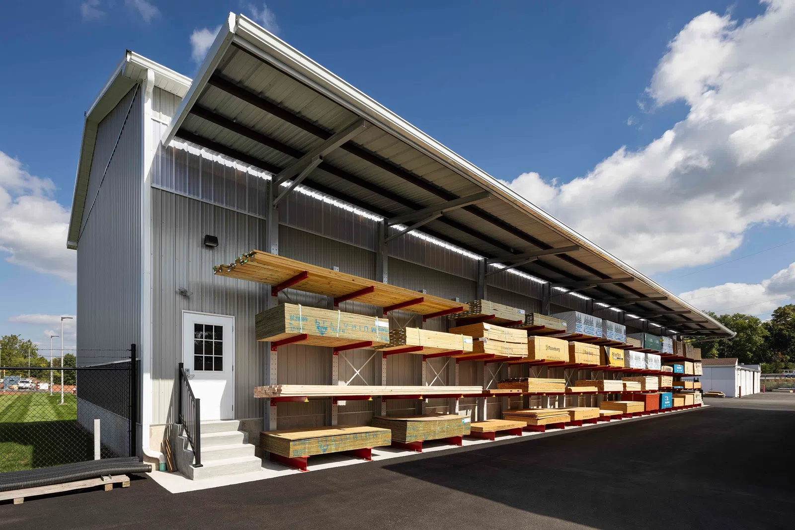 Outdoor lumber storage racks under metal roof with various wood planks on a sunny day with blue sky and clouds.