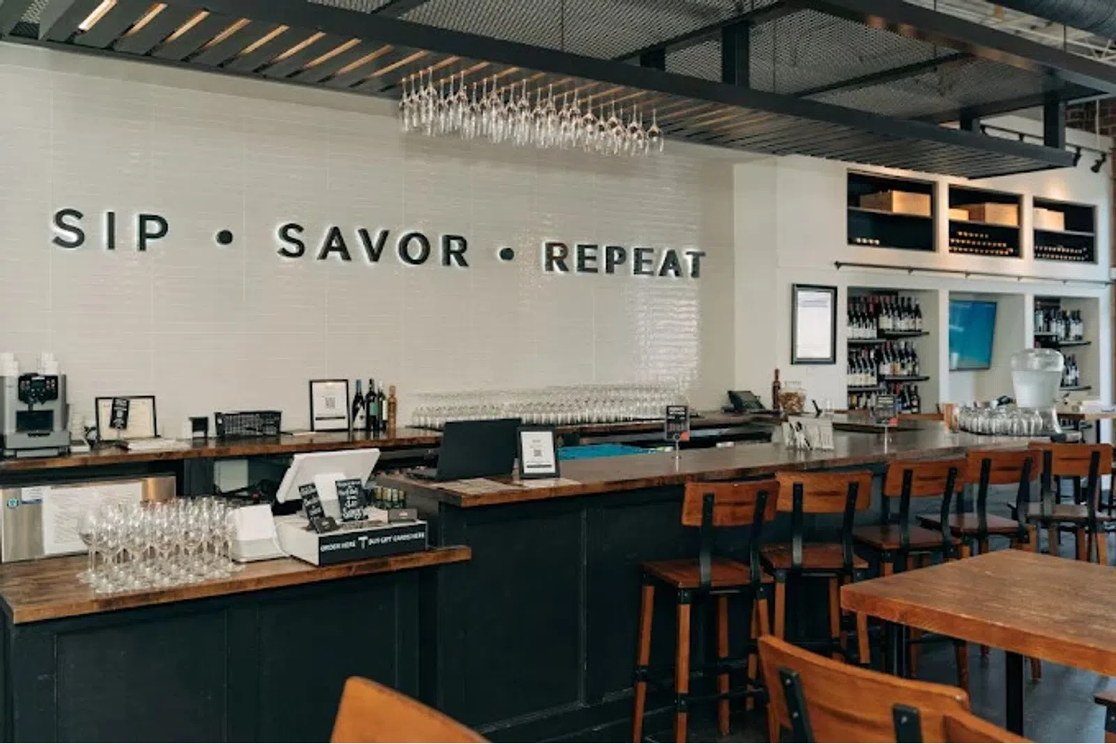 Modern bar interior with wooden stools, wine glasses, and wall text reading SIP SAVOR REPEAT above counter.