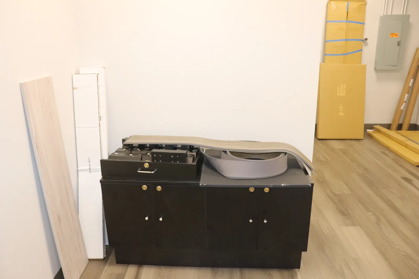 Black printer or copier machine in a room with wooden floor and white walls, surrounded by stacked boxes and panels