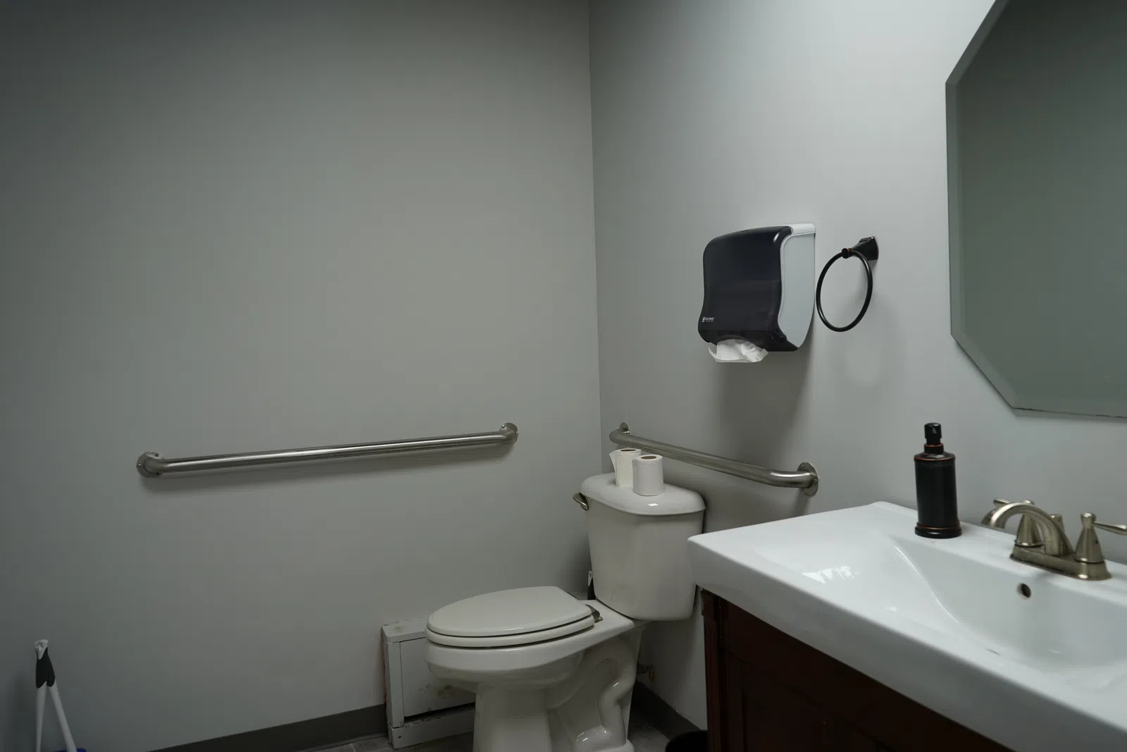 Accessible public restroom with toilet, sink, handrails, paper towel dispenser, and mirror in a clean, simple layout.