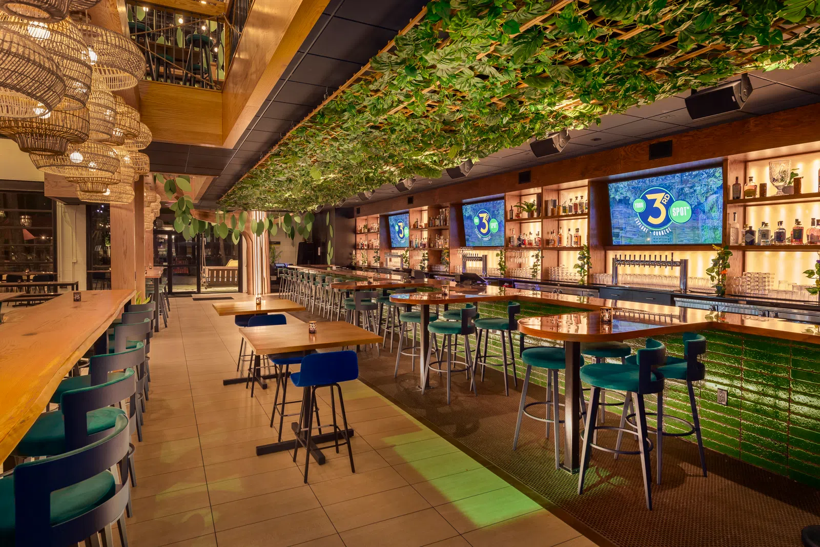 Modern bar interior with green ceiling plants, wooden tables, blue chairs, and illuminated shelves with bottles.