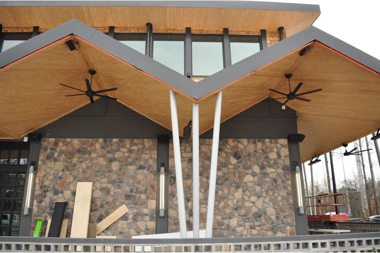 Modern building entrance with unique angular wooden roof, stone wall, ceiling fans, and metal support columns.