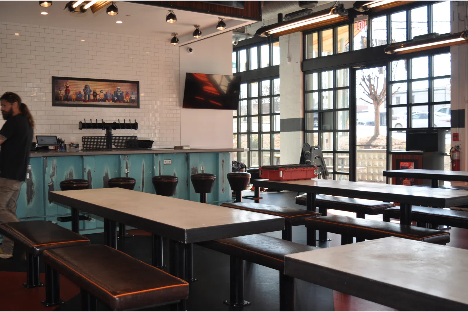 Modern indoor bar with long tables, leather benches, bar stools, large windows, and industrial lighting fixtures.