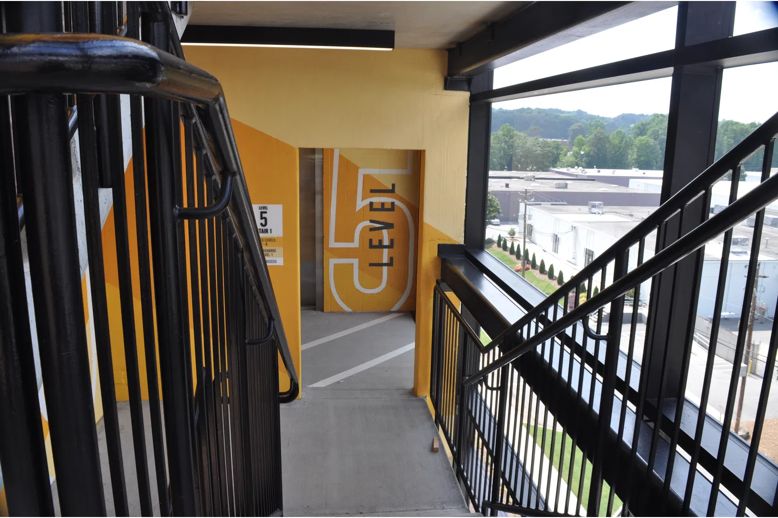 Staircase leading to level 5 of a parking garage with yellow walls and large windows overlooking an industrial area.