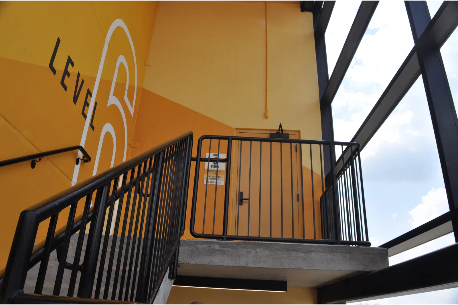 Stairwell leading to Level 6 with yellow walls, black railings, and large windows showing blue sky and clouds.