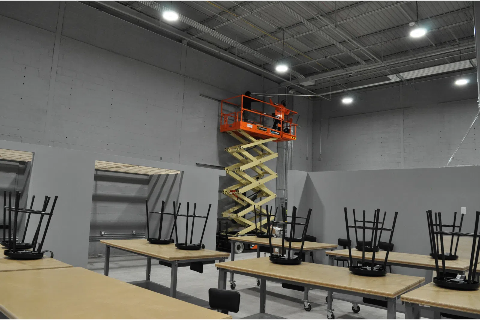 Scissor lift elevating two workers inside a large industrial room with tables and chairs stacked upside down.