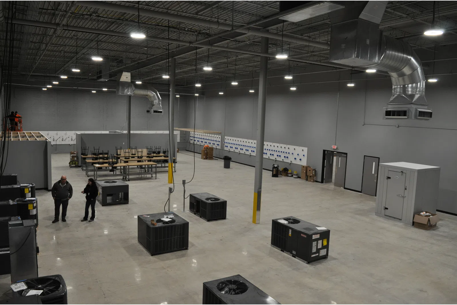 Spacious industrial warehouse with HVAC units on floor, high ceilings, ventilation ducts, two people and tables in background