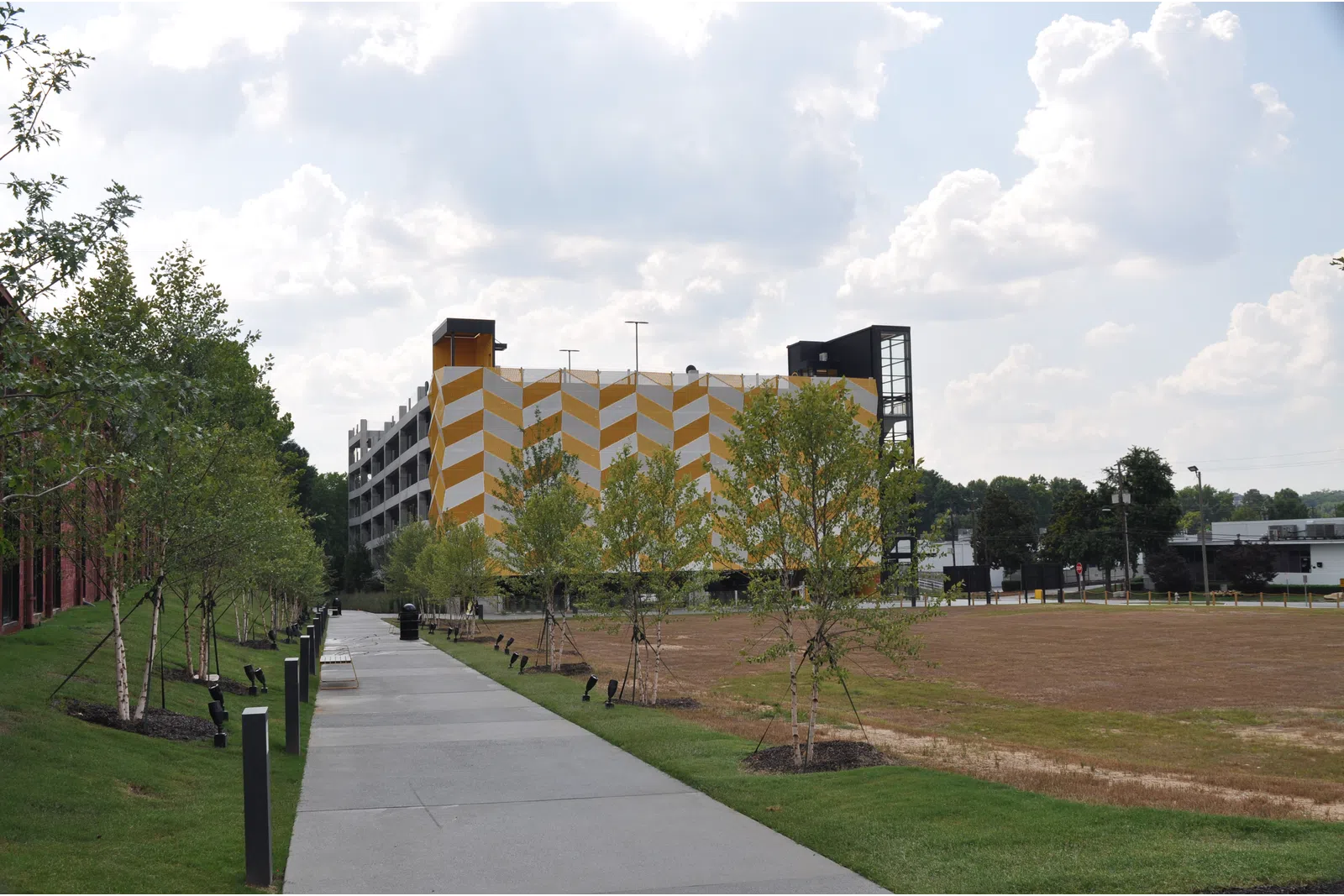 Pathway lined with young trees and lights next to a yellow and white patterned building under a cloudy sky.