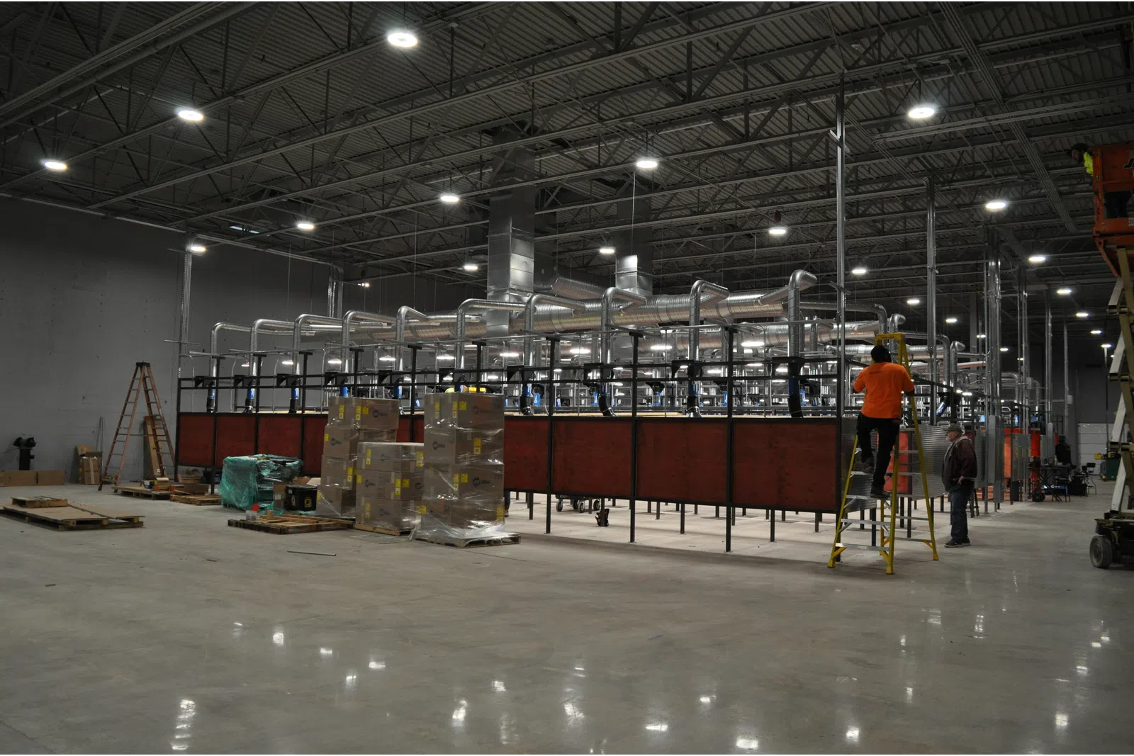 Large industrial warehouse with ventilation system, packed boxes, and workers installing equipment on a ladder.
