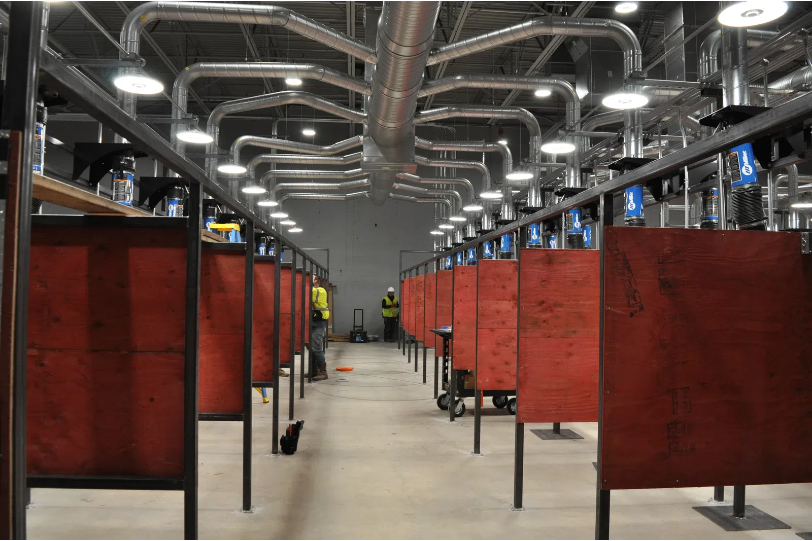 Indoor industrial workshop with rows of red wooden workstations and overhead ventilation pipes in a large space.