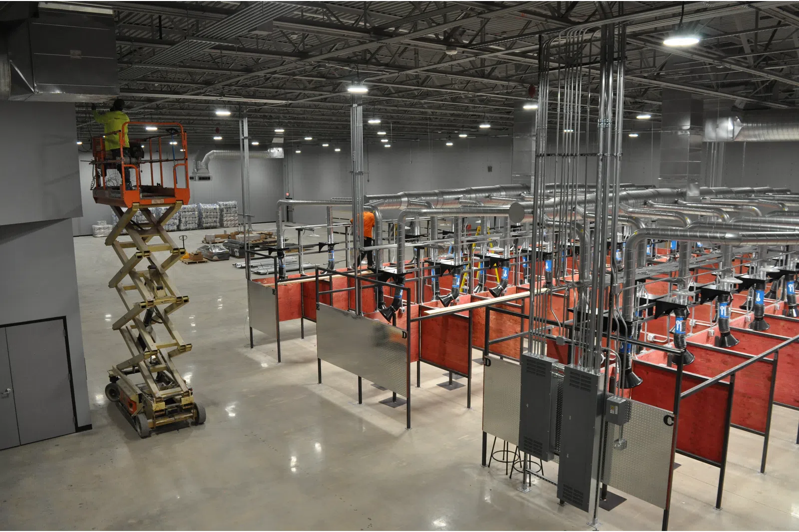 Worker on scissor lift in a large industrial welding training facility with ventilation and red partitions.