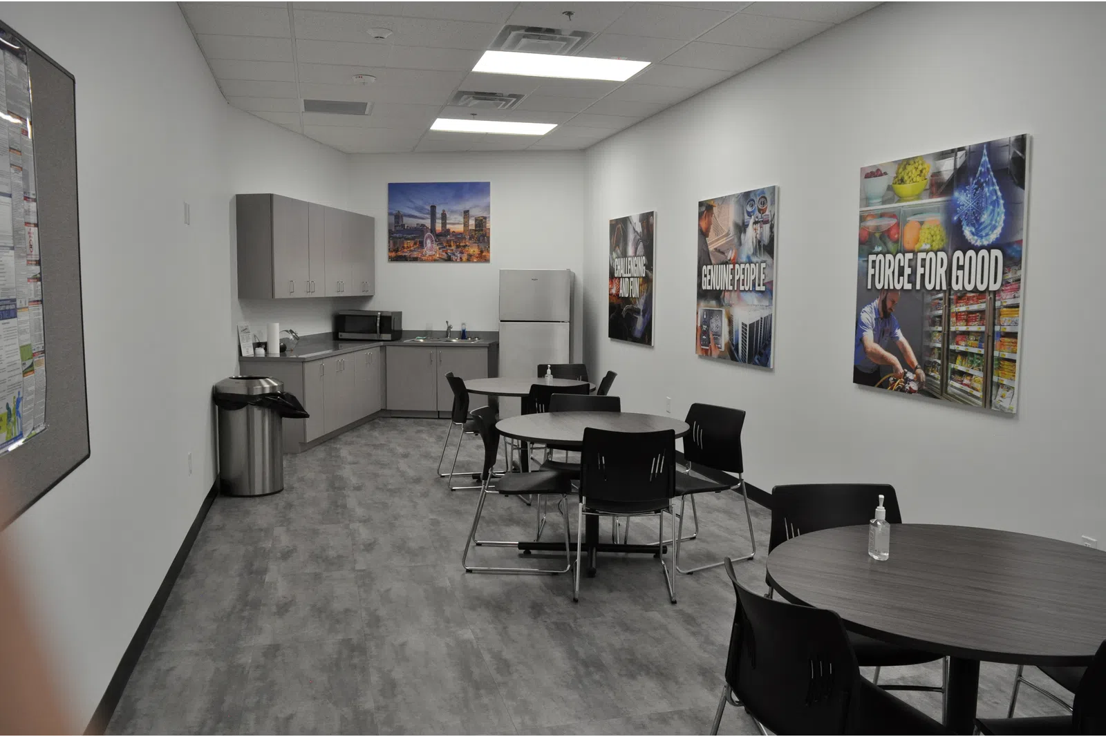 Corporate break room with round tables, black chairs, kitchenette, refrigerator, and motivational posters on walls.