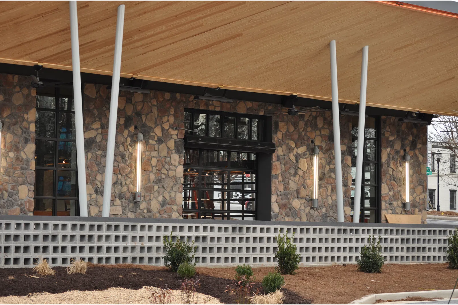 Modern building exterior with stone wall, large windows, vertical lights, and wooden roof overhang.