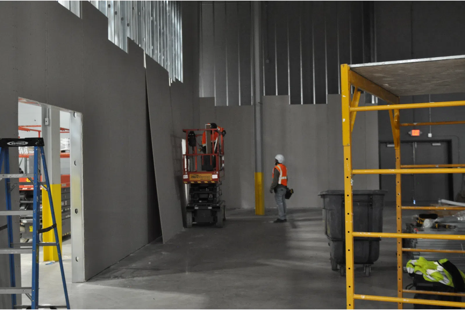 Construction worker in safety vest and helmet inside large unfinished industrial building with scaffolding and equipment.