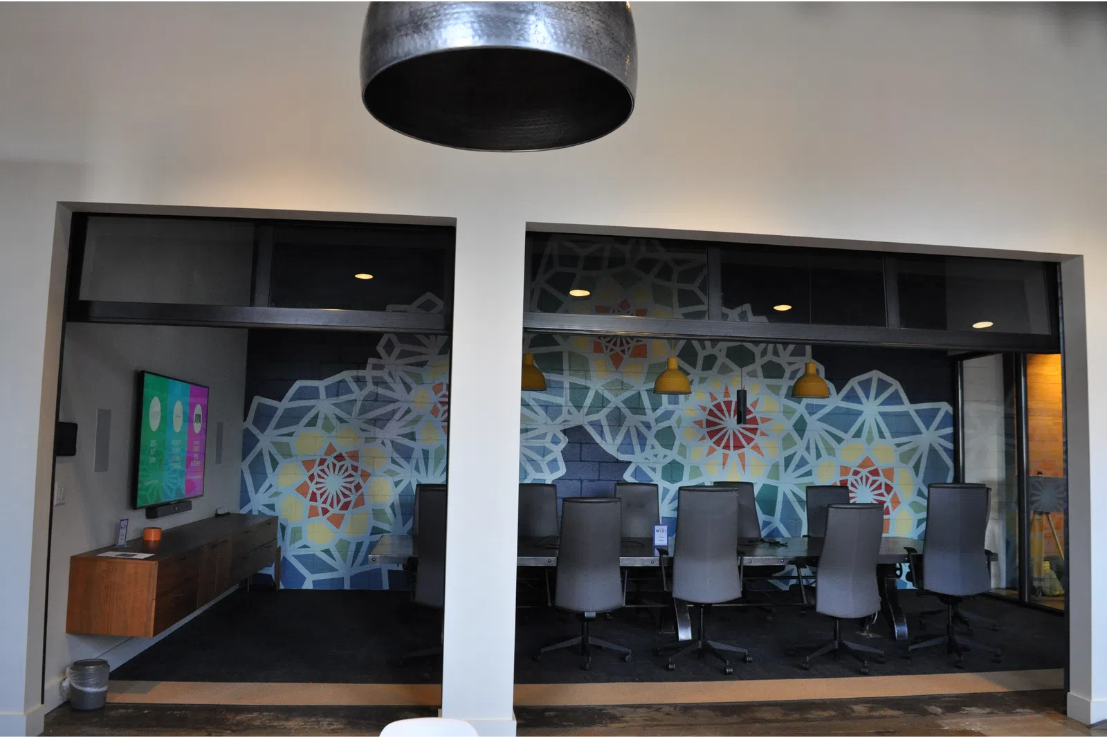 Modern conference room with patterned wall art, black chairs, pendant lights, and a mounted flat screen TV.