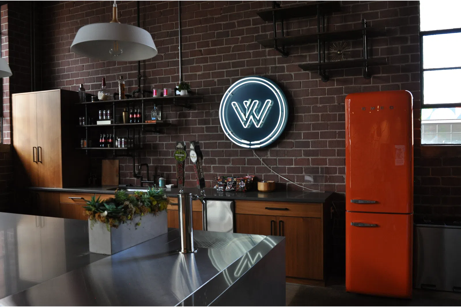 Industrial style kitchen with brick walls, neon sign, orange retro fridge, wooden cabinets, and modern stainless steel island.