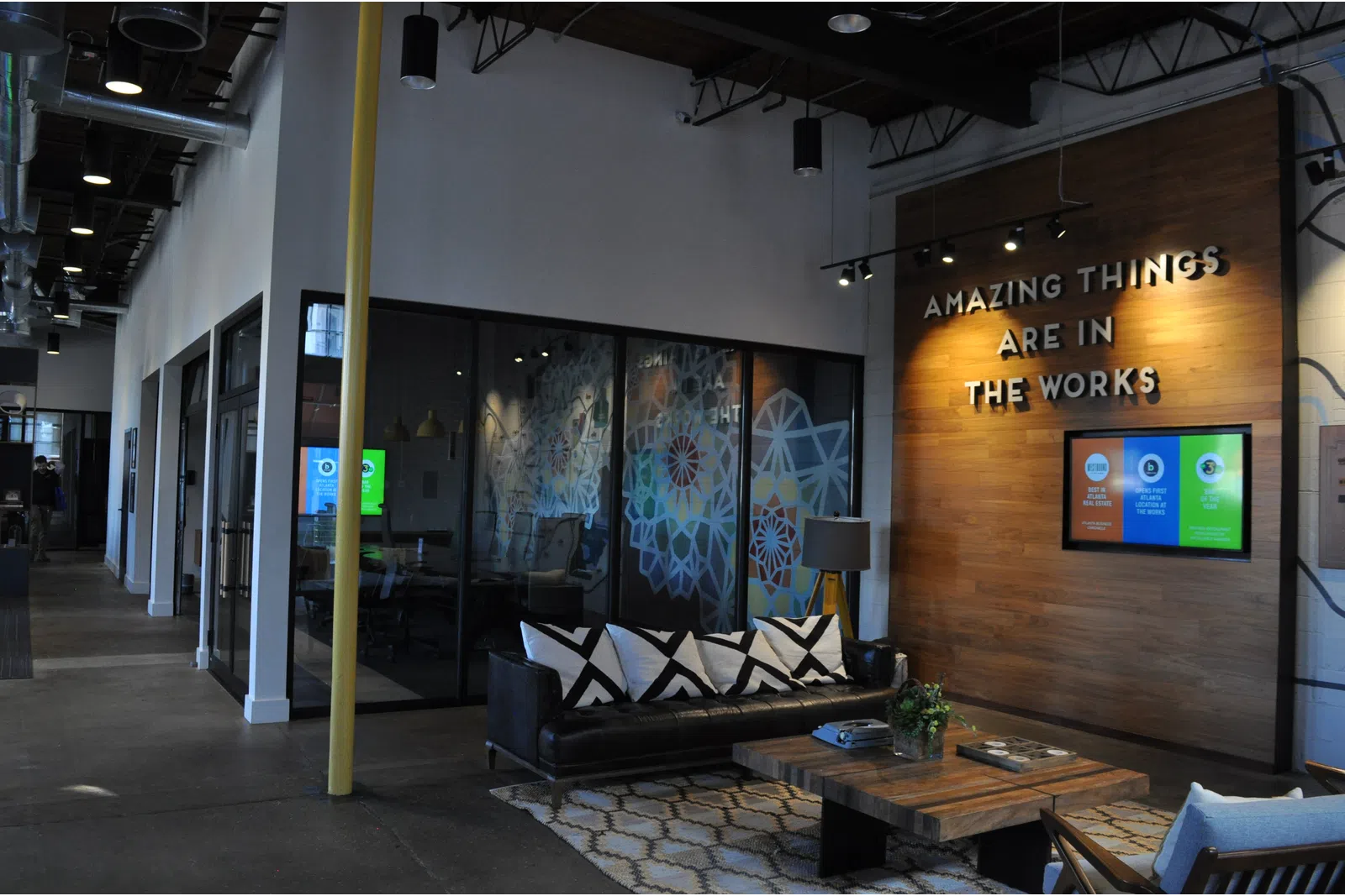 Modern office lounge with black sofa, wooden coffee table, inspirational wall sign, and glass meeting rooms.