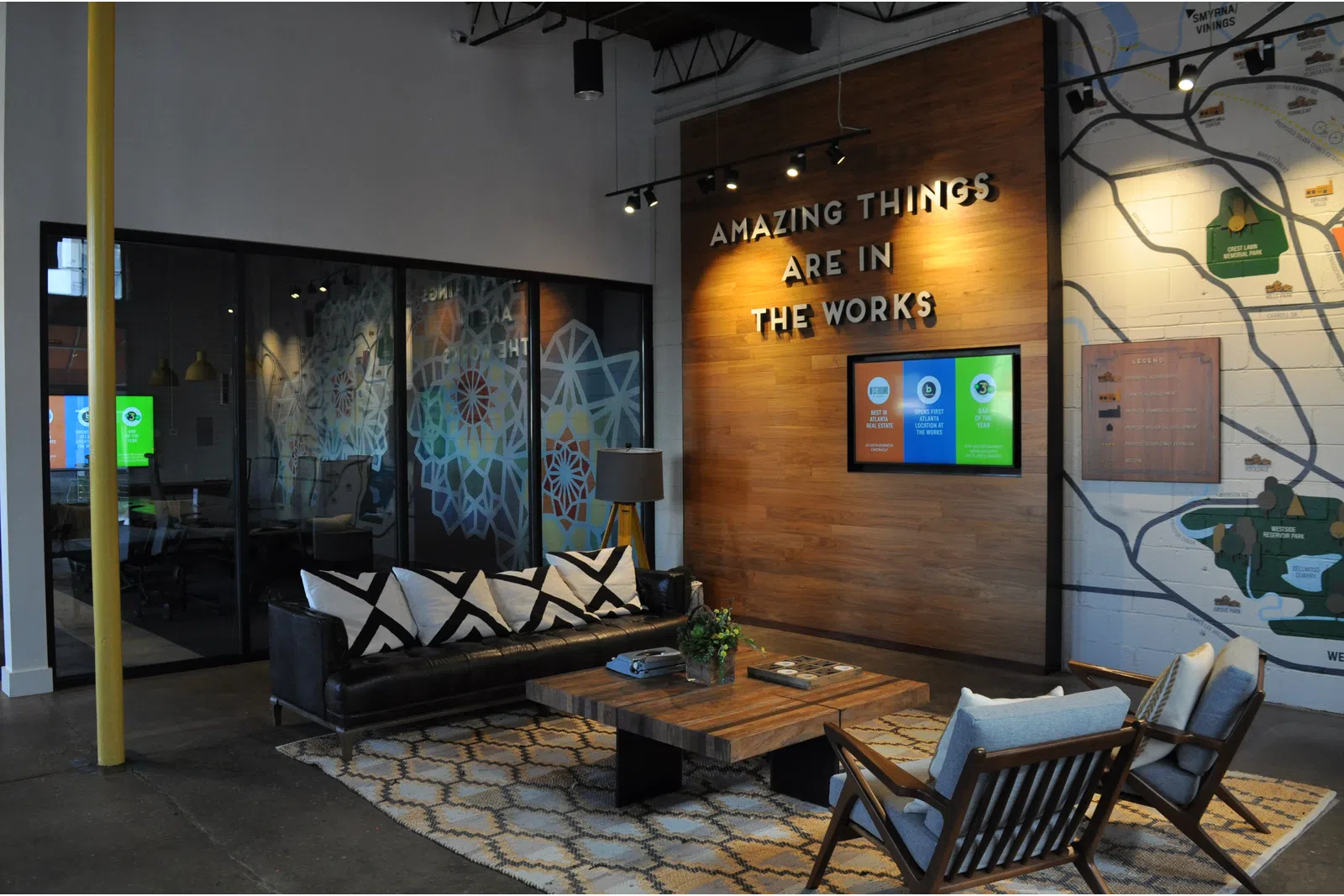 Modern office lounge with black couch, wooden table, patterned rug, wall sign reading Amazing Things Are In The Works