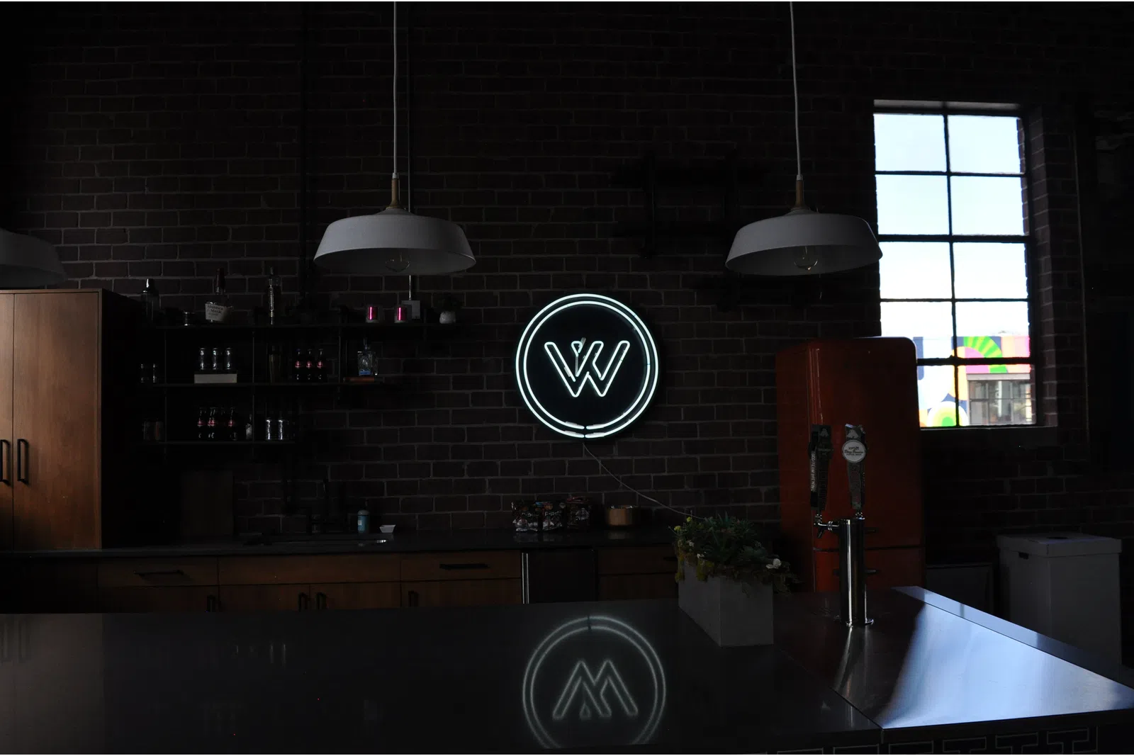 Dark industrial kitchen with brick walls, hanging lights, neon W logo, and orange refrigerator by a window.