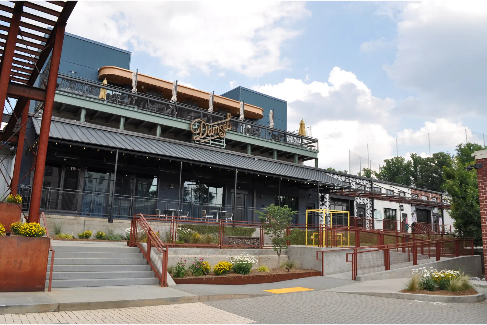 Modern restaurant building with outdoor seating, stairs, ramps, and flower beds under partly cloudy sky.