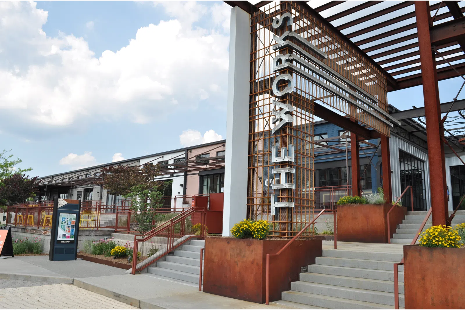 Exterior of a modern building with rust-colored metal structure and stairs under a cloudy sky.
