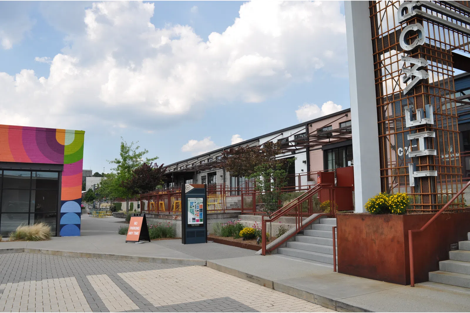 Outdoor urban plaza with colorful mural, modern buildings, stairway, and clear sky with scattered clouds.