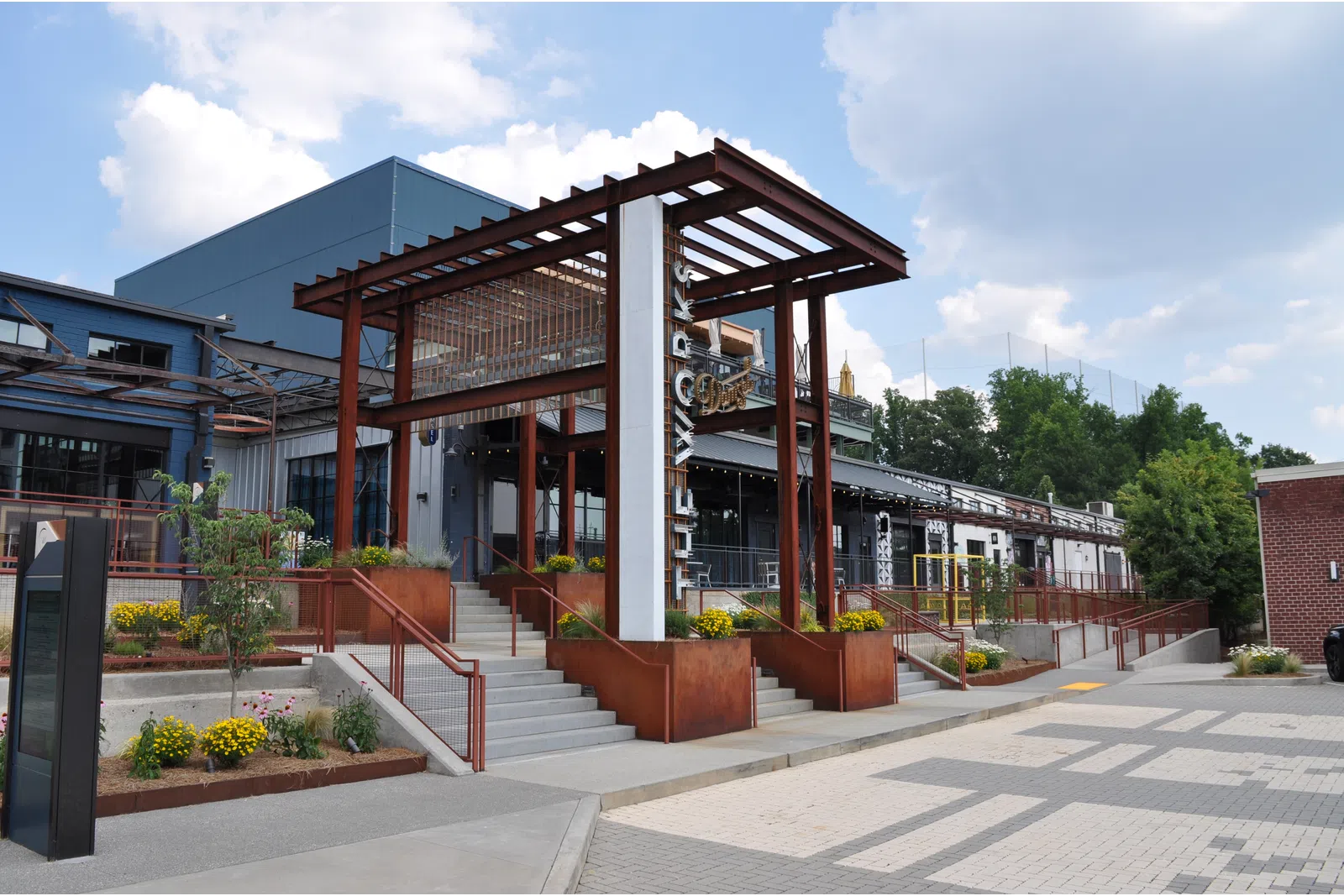 Modern building entrance with rust-colored steel pergola, concrete stairs, and landscaped planters under a partly cloudy sky