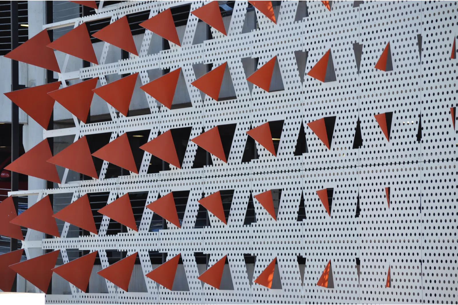 Facade with rows of red triangular metal elements mounted on perforated white panels of a building exterior.