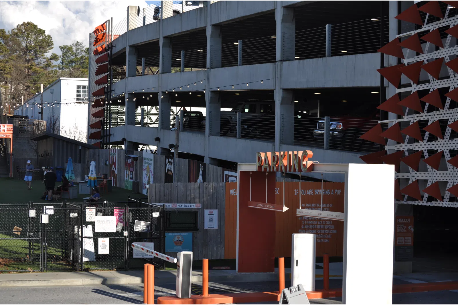Outdoor parking garage entrance with orange signage and nearby green fenced area under partly cloudy sky.