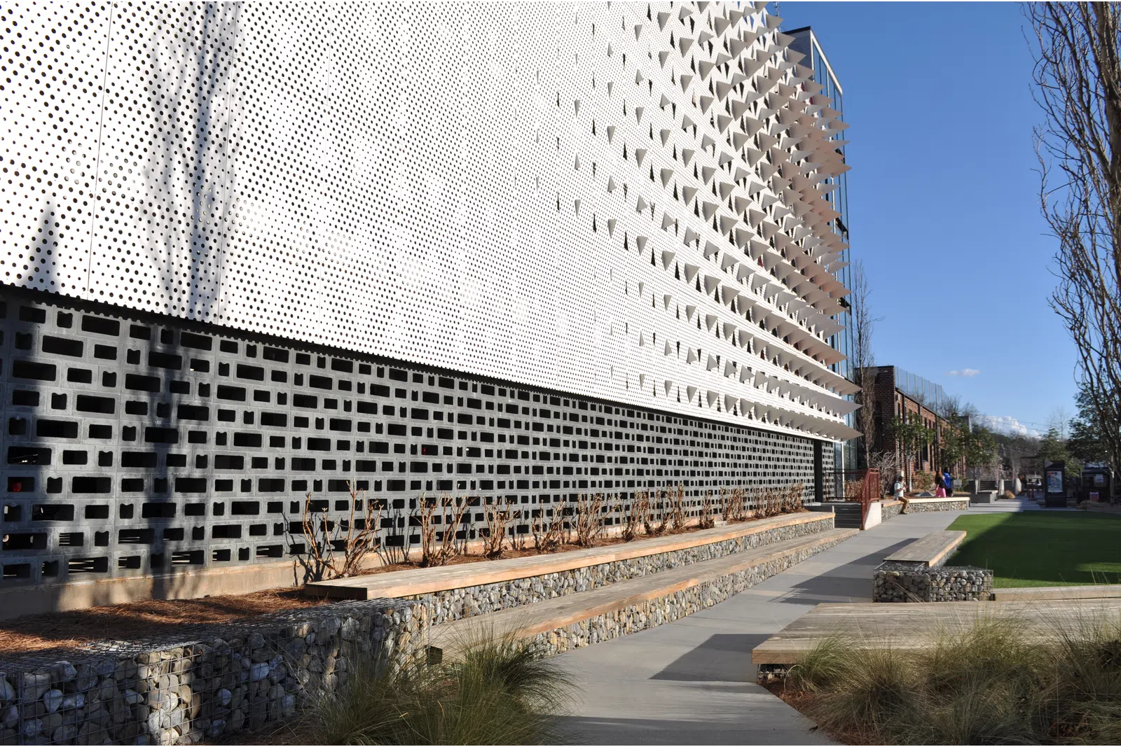 Modern building facade with perforated white panels, geometric patterns, and landscaped outdoor seating area under clear sky.