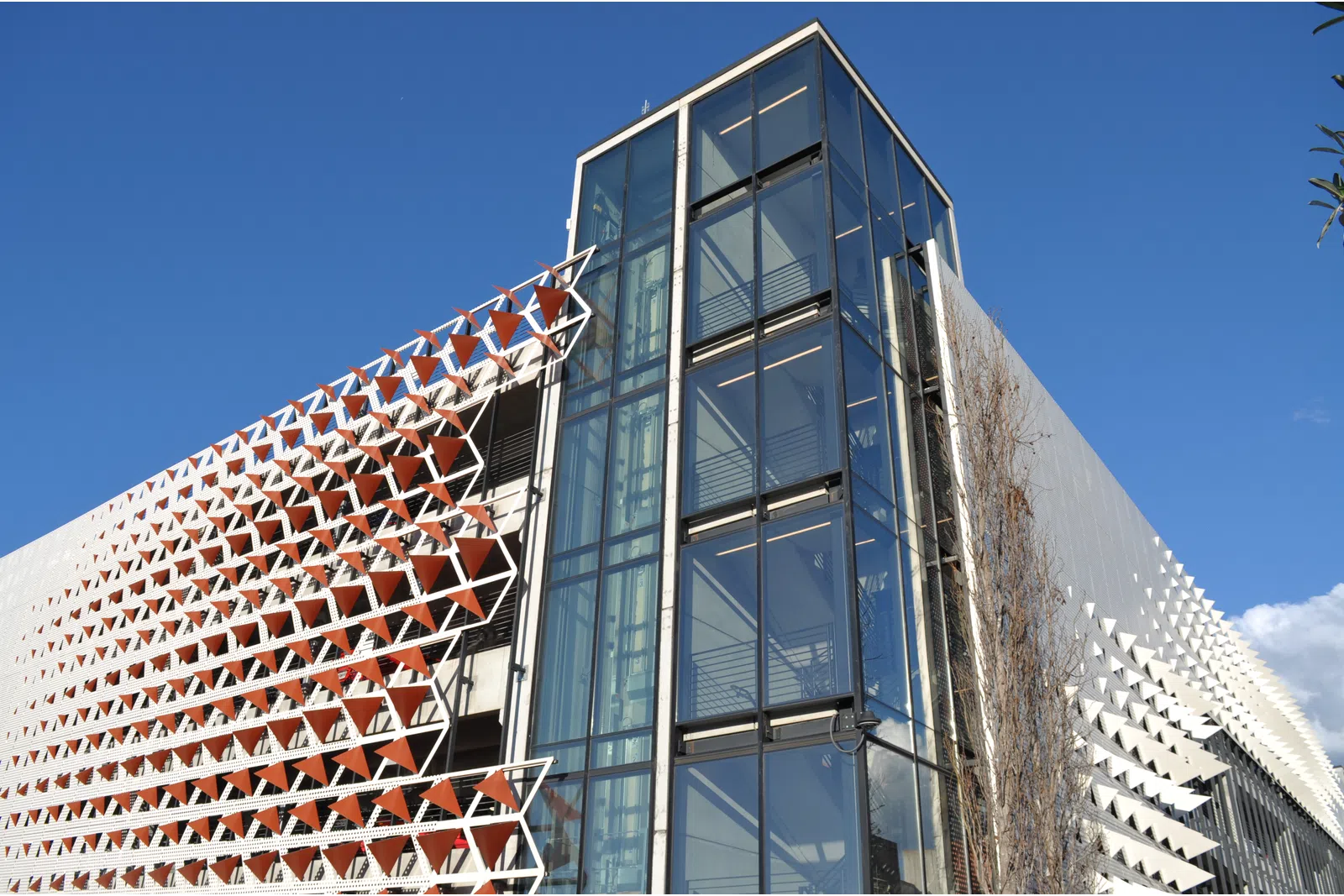 Modern building facade with geometric sunshades and glass elevator tower under clear blue sky