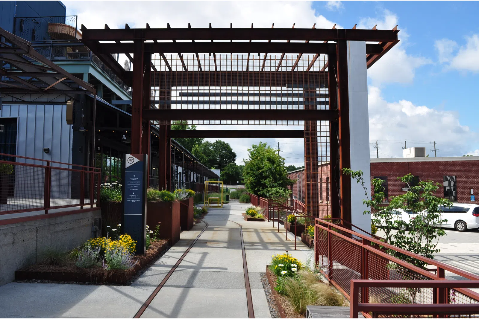 Modern outdoor walkway with metal pergola, planters, and urban buildings under a blue sky with clouds.