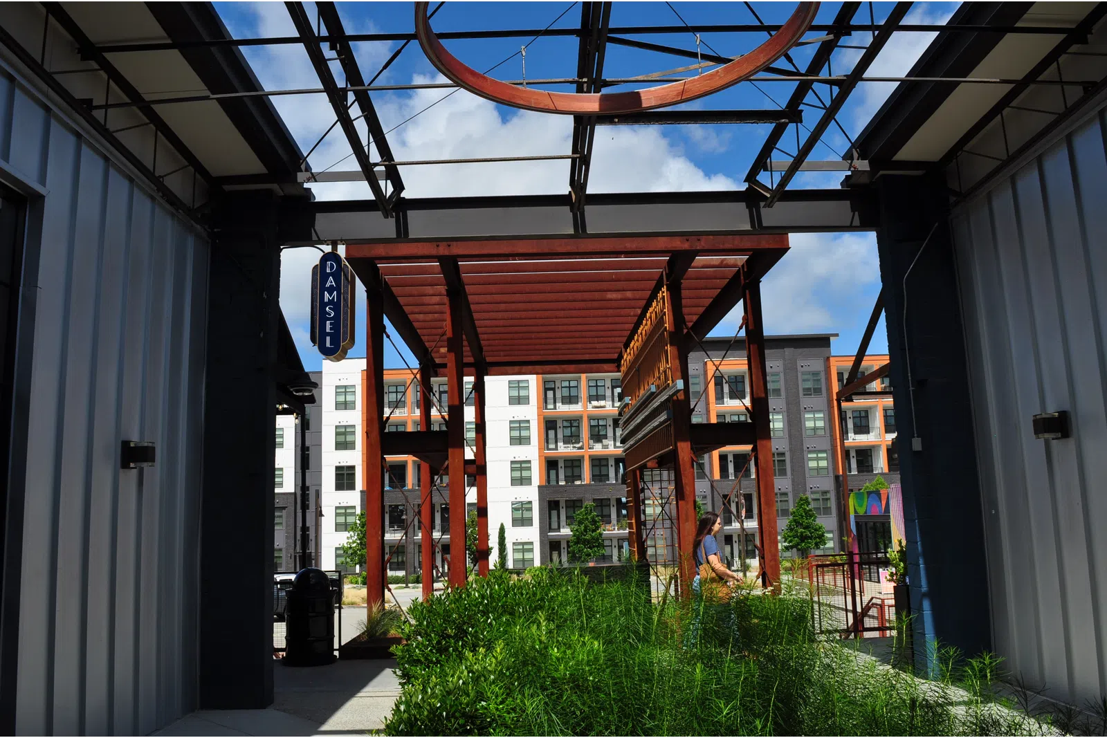 Outdoor modern urban area with metal structures, greenery, and a person walking near apartment buildings on a sunny day
