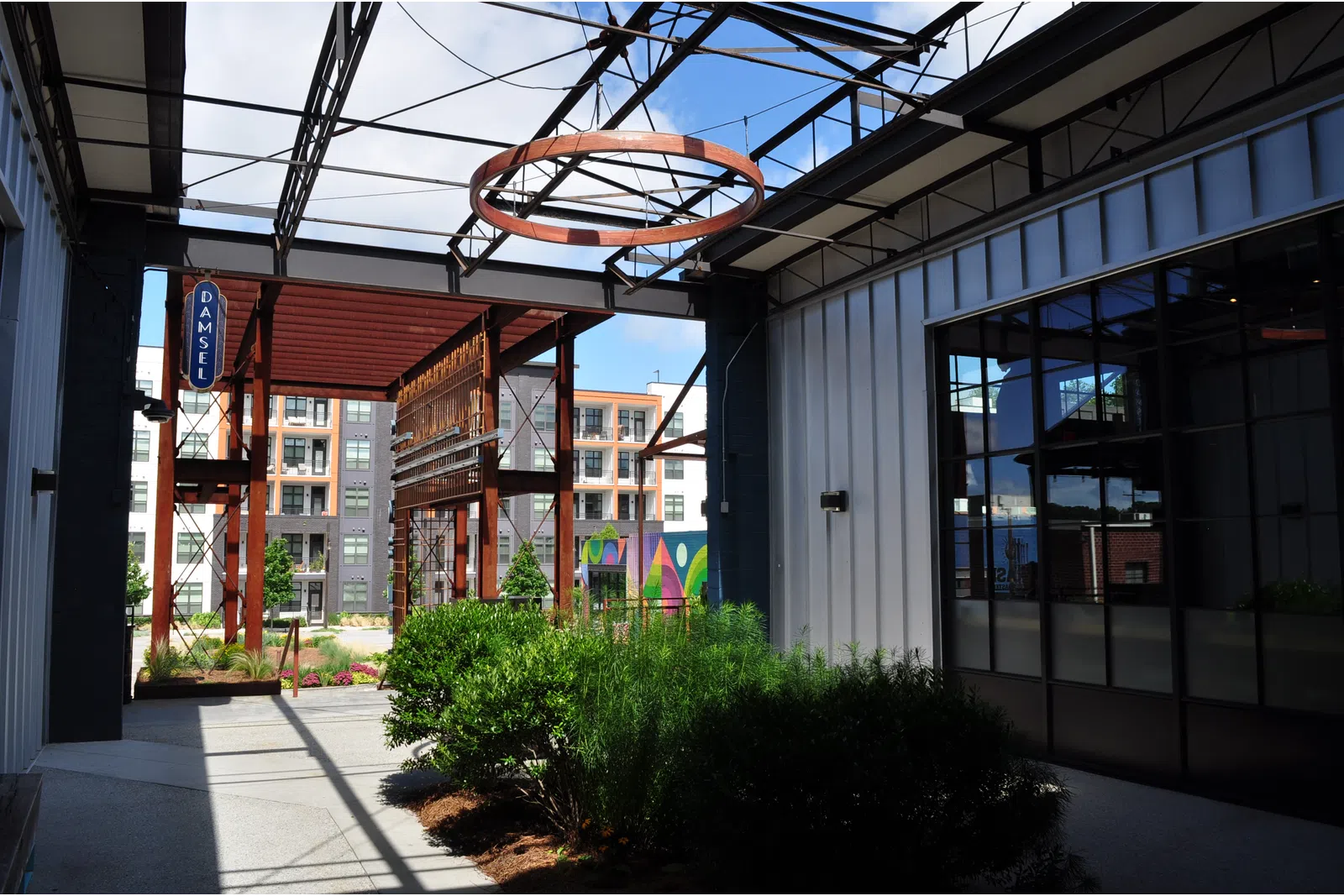 Modern industrial-style outdoor corridor with greenery, metal framework, glass windows, and apartment buildings in background
