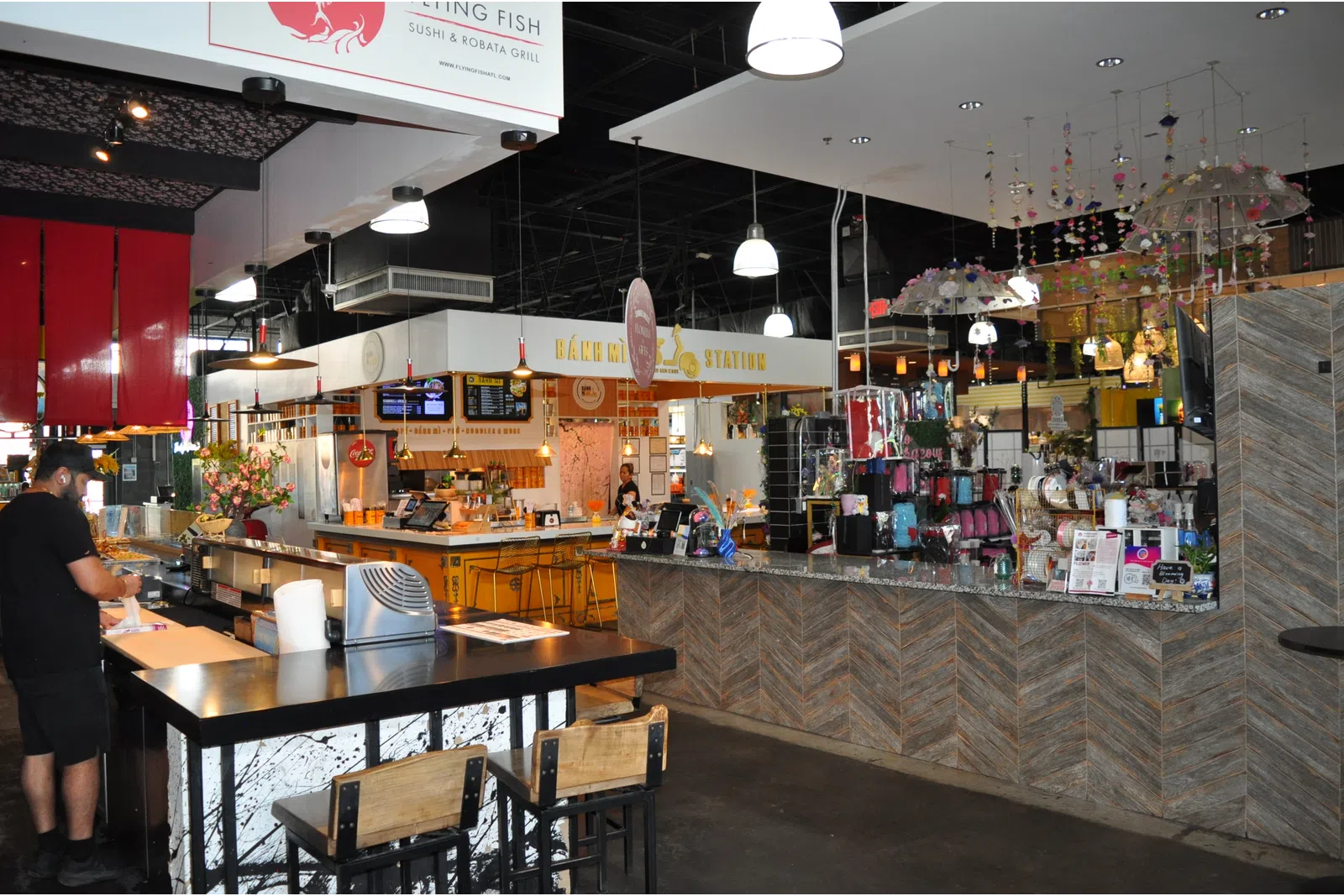 Modern food court interior with sushi bar, colorful decor, hanging lamps, and seating areas with customers.