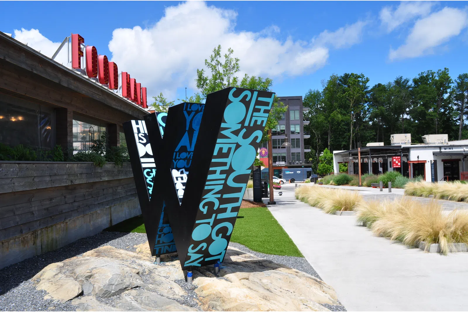 Large decorative 'W' sculpture with text in an outdoor urban park area under blue sky.