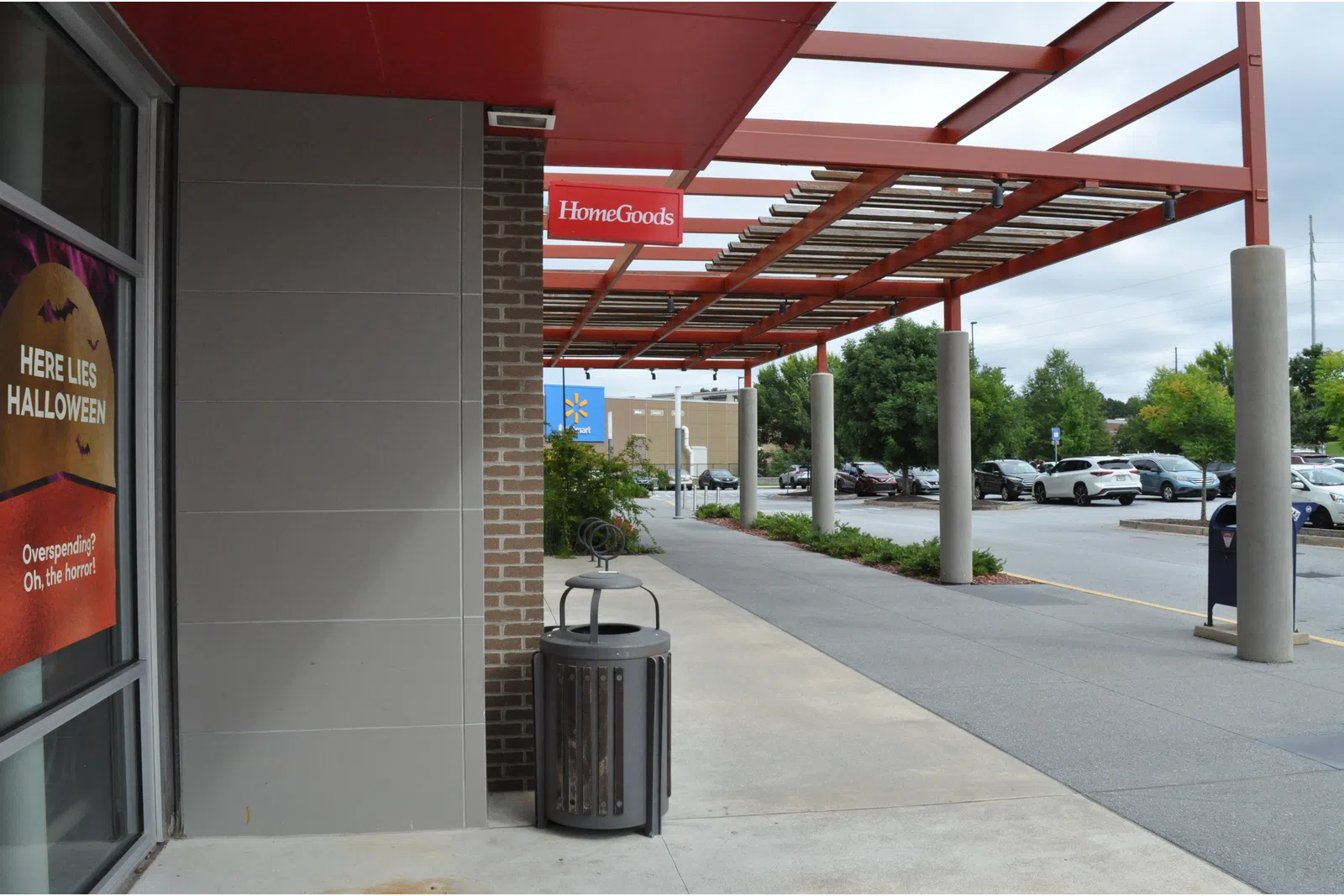 Outdoor shopping area with HomeGoods sign, parking lot, trash can, and Halloween sale advertisement on storefront window.