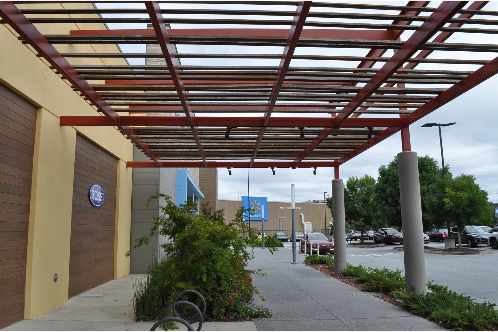 Outdoor shopping area with Ross and Walmart stores, covered walkway, plants, and parked cars on a cloudy day