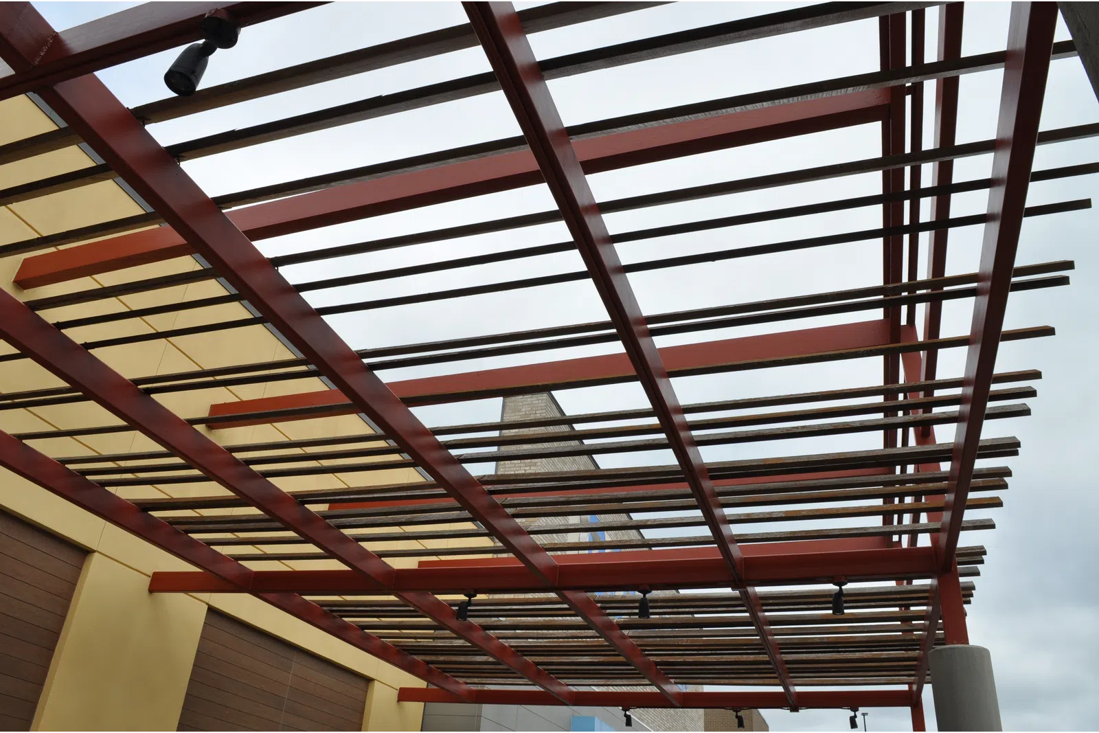 Modern pergola with wooden slats and red beams over a building exterior under cloudy sky.