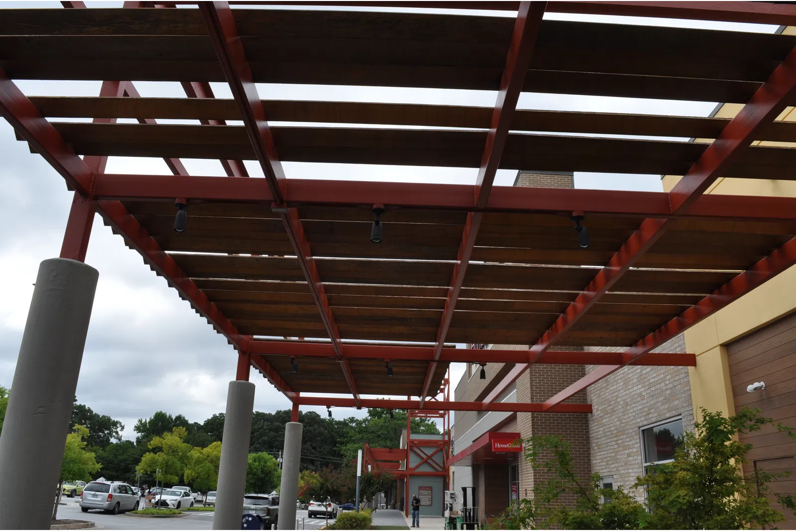 Outdoor walkway with red steel pergola and wooden slats next to a brick building under cloudy sky.