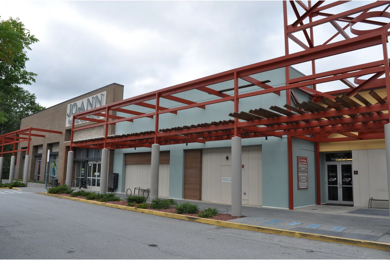 Exterior view of a shopping center with Jo-Ann fabric store and covered walkway with red metal framework.